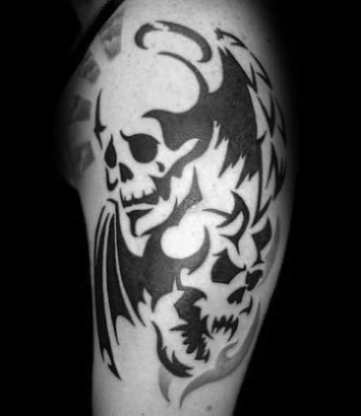 Bold tribal skull tattoo on upper arm with sharp black shapes, demonic features, and wing-like elements creating a fierce composition.