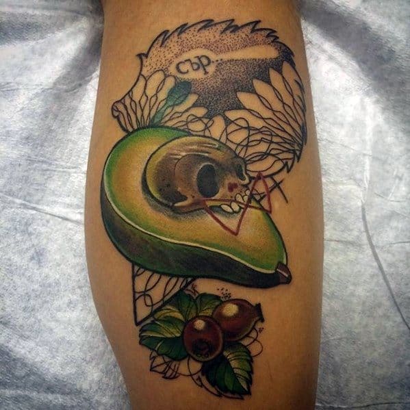 Detailed avocado tattoo with a skull pit, wings, and foliage, blending dark and botanical elements in a bold calf design.