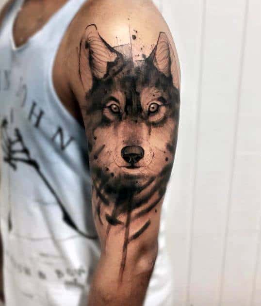 Realistic black and grey wolf tattoo on upper arm with bold shading, soft brushstroke edges, and intense, focused eyes.