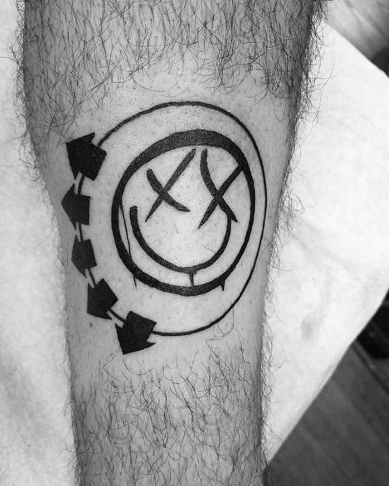 Black and white tattoo of the Blink-182 smiley face logo with bold black lines and arrows, placed on the forearm.