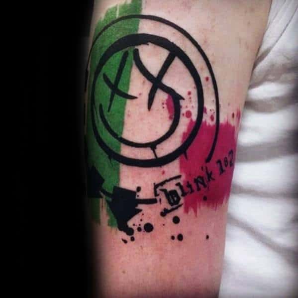 Tattoo of the Blink-182 smiley face logo with vibrant green and pink watercolor splashes, accompanied by the band name "Blink 182" in black ink, set against a bold, graphic design.