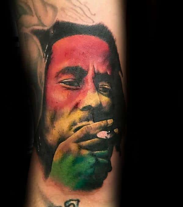 Color portrait tattoo of Bob Marley smoking, with red, yellow, and green hues blending across his face in a bold Rasta-inspired design.
