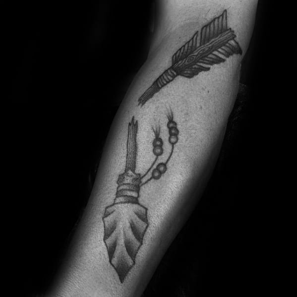 Black and grey broken arrow tattoo with a leaf-shaped arrowhead and bead details, split cleanly in two across the forearm.