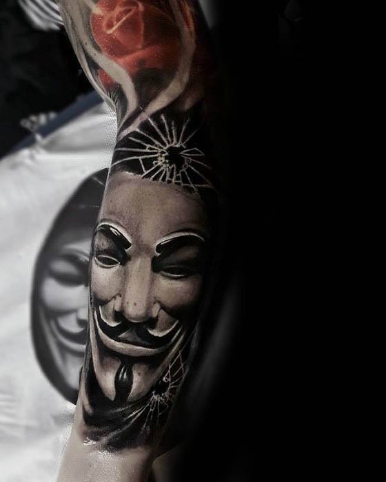 Tattoo of a Guy Fawkes mask beneath shattered glass, blending rebellion symbolism with bold black and grey realism on the forearm.