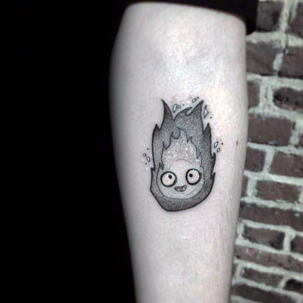 Black and grey Calcifer tattoo with detailed dotwork shading, playful eyes, and flickering flame tips on the forearm.