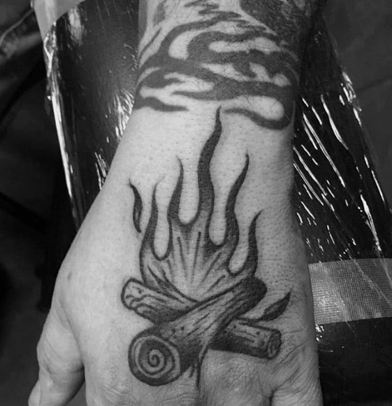 Black and grey campfire tattoo on the hand with crossed logs and stylized flames in a classic illustrative style.