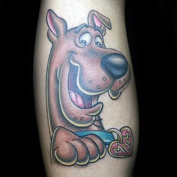 Cheerful Scooby-Doo tattoo with a heart-shaped dog tag that says “Mom,” showing his playful smile and friendly expression on the leg.