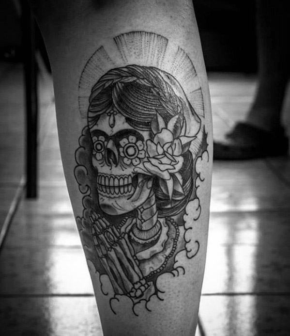 La Catrina tattoo of a praying skeleton woman with floral hairpiece and halo rays, designed in bold black linework and shading.