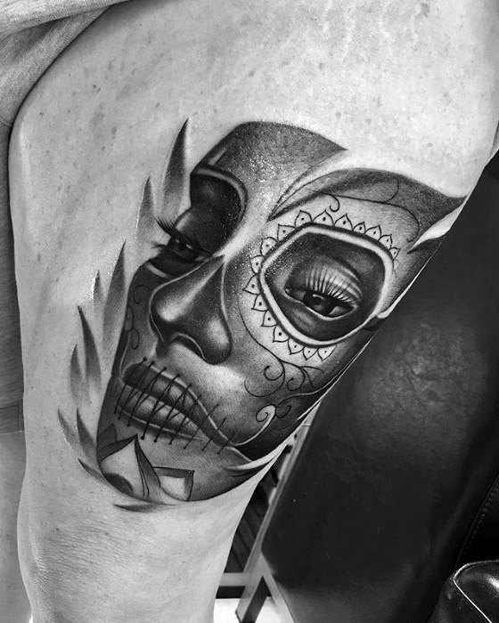La Catrina tattoo of a woman's face in sugar skull makeup with bold shading and sharp abstract elements on the thigh.