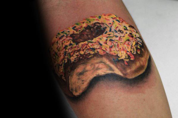 Realistic tattoo of a bitten chocolate-covered donut with colorful sprinkles, featuring detailed shading and texture for a lifelike appearance.