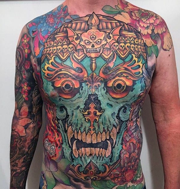Full chest Tibetan skull tattoo with turquoise face, golden crown, fiery details, and vivid floral elements extending to both arms.