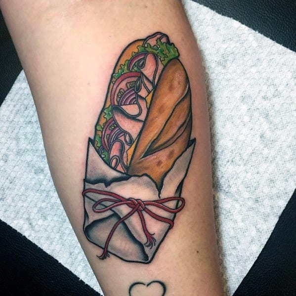 Colorful tattoo of a baguette sandwich wrapped in paper and tied with a red string, featuring detailed layers of meat, lettuce, and onions.