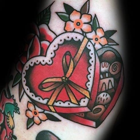 Traditional-style tattoo of a heart-shaped chocolate box with a golden ribbon, surrounded by flowers, featuring bold lines and vibrant colors.