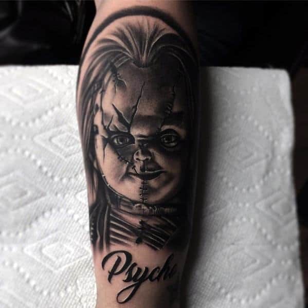 Black and grey Chucky tattoo with deep shading, featuring his eerie grin, stitched face, and the word "Psycho" for a bold horror look.