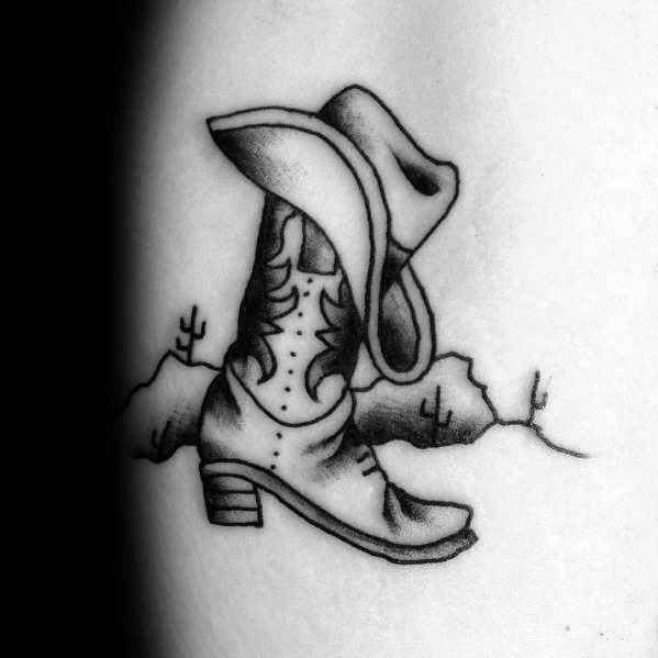 Black and white tattoo of a cowboy boot with a hat resting on top, set against a desert backdrop with cacti, symbolizing Western heritage.