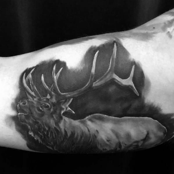 Realistic black and grey elk tattoo on the bicep, featuring detailed shading, bold antlers, and a misty background for a lifelike design.