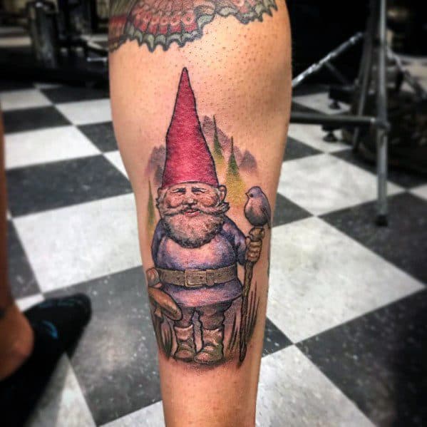 Tattoo of a friendly gnome with a red hat and blue coat, holding a wooden staff with a small bird perched on it, set against a forest backdrop.