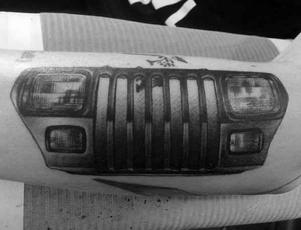 Realistic black and grey Jeep tattoo on the forearm, featuring a detailed front grille and headlights, creating a bold and rugged design.