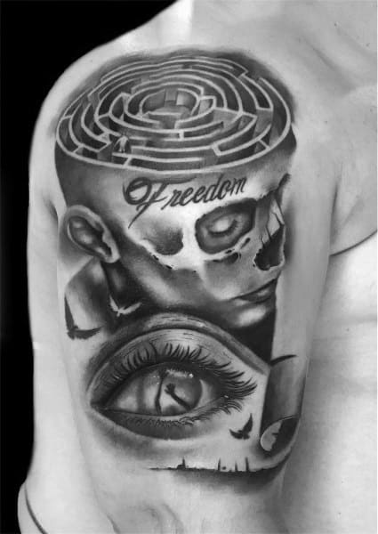 Surreal black and grey tattoo on a man's shoulder, featuring a maze inside a skull, the word "Freedom," and a realistic eye design.