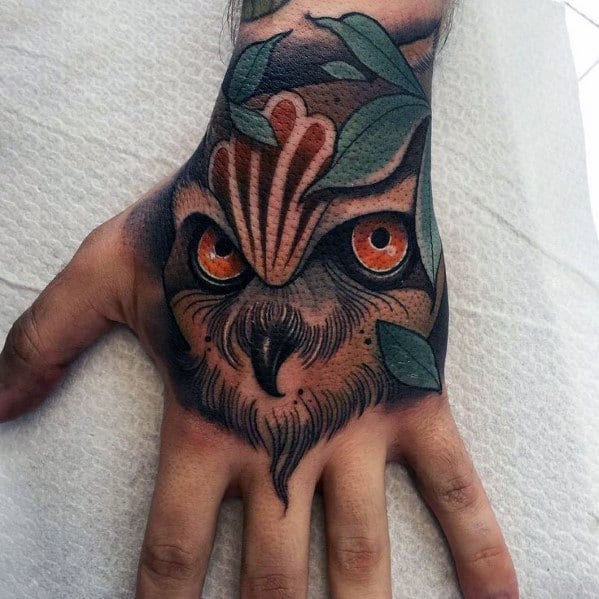 Neo-traditional owl hand tattoo with piercing orange eyes, bold shading, and leaf accents, blending nature-inspired elements with striking details.