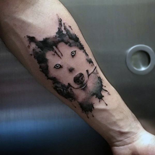 Watercolor-style Siberian Husky tattoo on a man's forearm, featuring bold splashes of black ink and piercing eyes for a striking effect.