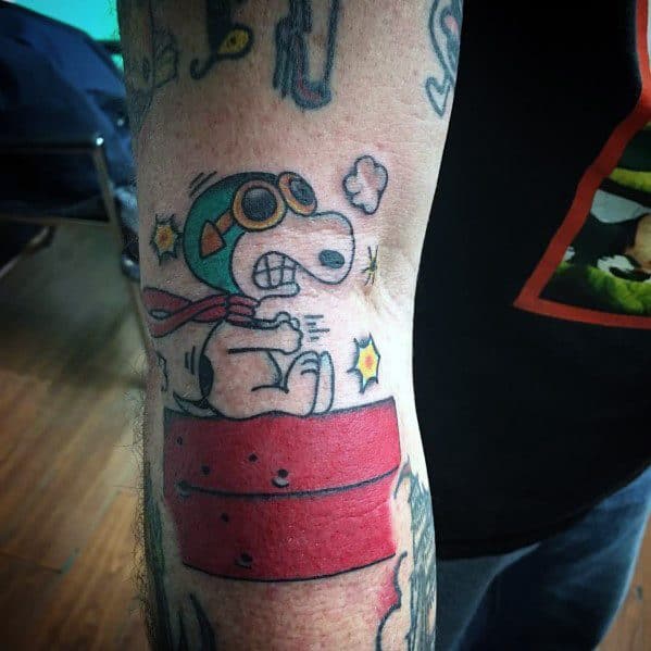 Colorful tattoo of Snoopy as a flying ace, wearing goggles and a scarf, sitting on a red doghouse with action lines and stars.