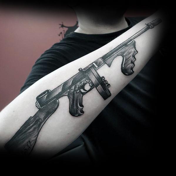 Realistic black and grey Tommy gun tattoo on the forearm, featuring intricate shading and fine details for a bold and striking firearm design.