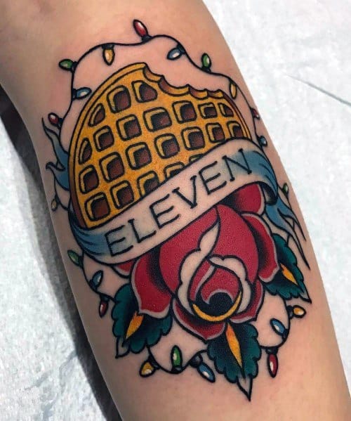 Traditional-style waffle tattoo with a banner reading "Eleven," a red rose, and colorful Christmas lights, inspired by pop culture and vintage designs.