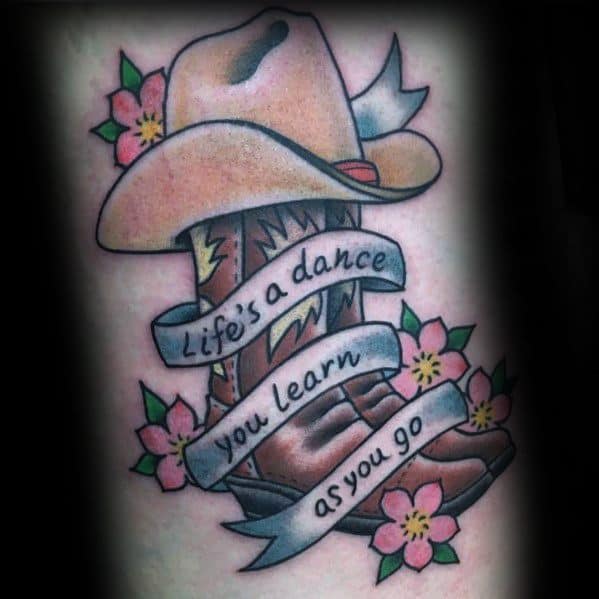 Tattoo of a cowboy boot and hat with a banner reading "Life’s a dance, you learn as you go," surrounded by flowers, symbolizing resilience and growth.