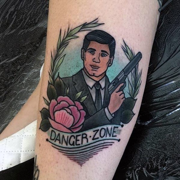 Archer tattoo featuring the spy in a suit holding a gun, framed by laurel leaves, a pink rose, and a 'Danger Zone' banner.
