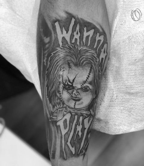 Black and grey Chucky tattoo featuring his stitched face, eerie grin, and the phrase "Wanna Play?" in bold lettering for a haunting horror design.