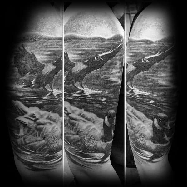 Realistic black and gray tattoo sleeve of geese swimming and taking flight over a reflective water surface, with intricate shading and fine details.