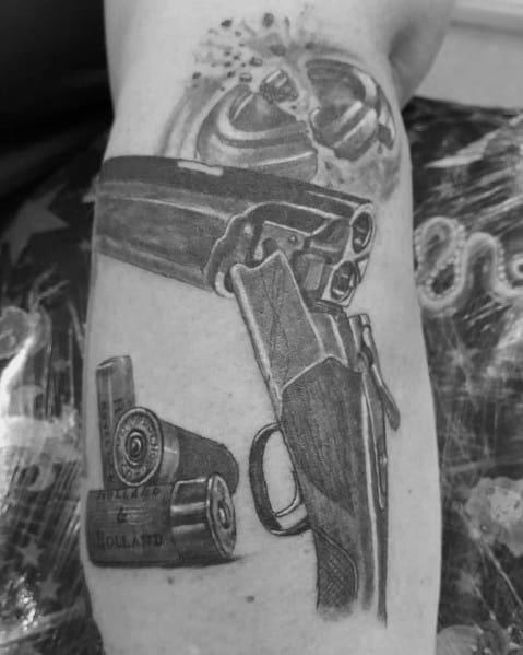 Realistic black and grey tattoo of a double-barrel shotgun with shotgun shells and a shattered clay target, showcasing fine shading and detail.