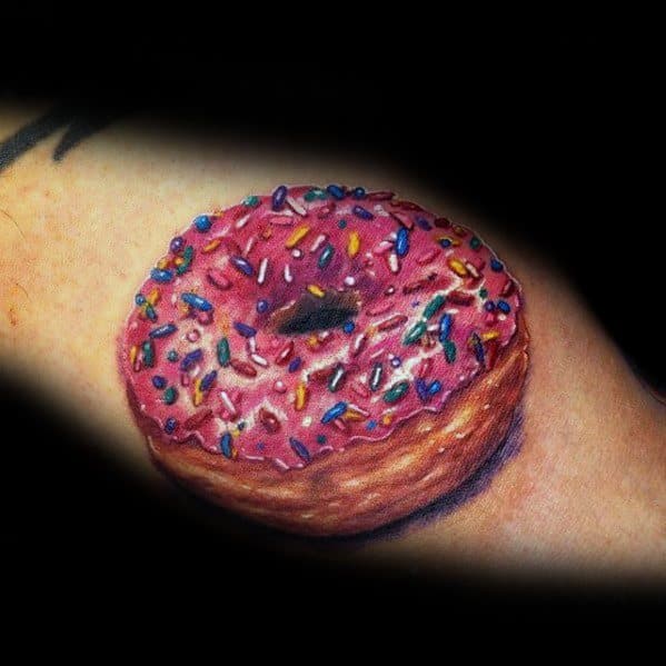 Tattoo of a hyper-realistic pink frosted donut with vibrant sprinkles, detailed shading, and a 3D effect on the skin.