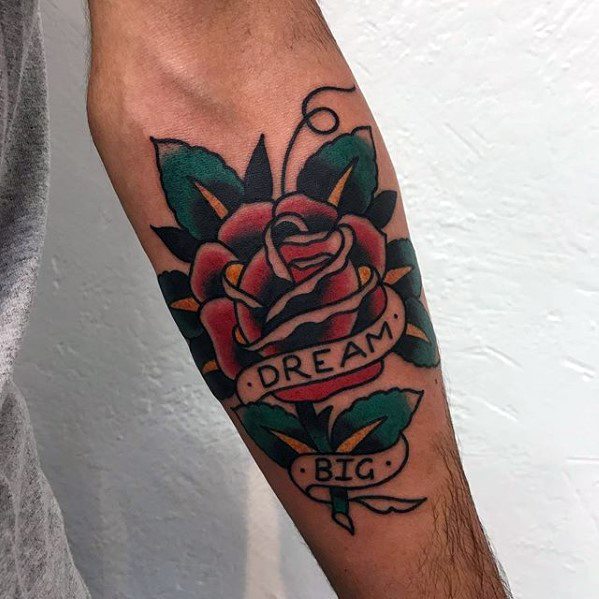 Forearm tattoo of a traditional red rose with green leaves, wrapped in a banner that reads “DREAM BIG” in bold capital letters.