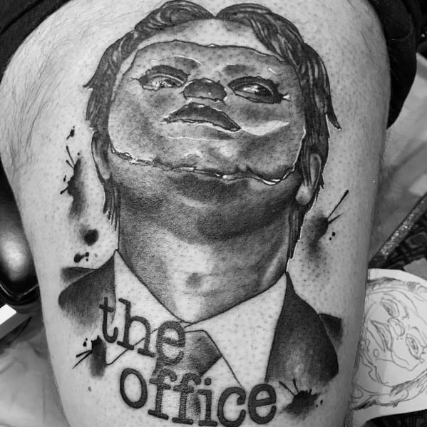 Black and grey tattoo of Dwight Schrute with a puppet-like face, featuring bold shading and "The Office" text in a splattered ink style.