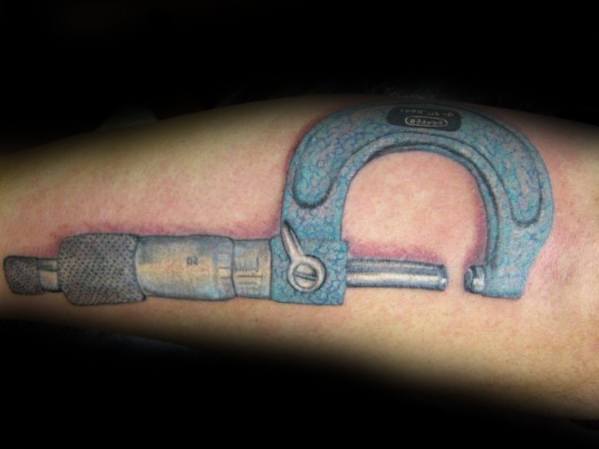 Tattoo of a realistic blue micrometer with detailed shading and texture, wrapped around the arm to resemble a precision tool embedded in the skin.
