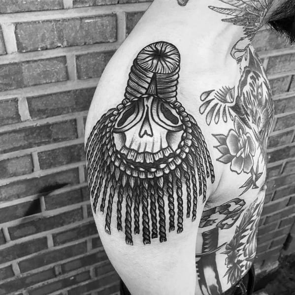 Striking epaulette tattoo with a sugar skull motif, thick braided ropes, and long tassels draping over the upper arm.