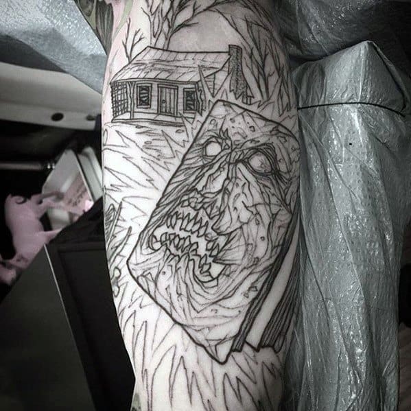 Black and grey linework Evil Dead tattoo featuring the Necronomicon with a twisted face below the haunted cabin in a creepy forest scene.