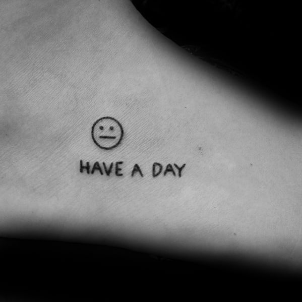 Minimalist tattoo of a neutral face emoji with the phrase "HAVE A DAY" on the skin