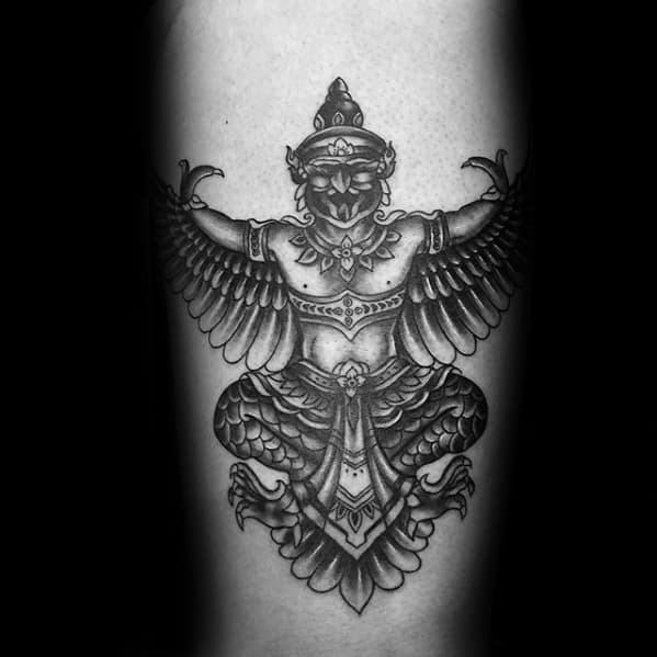 Garuda tattoo featuring a symmetrical winged figure with detailed feathers, ornate jewelry, and a calm expression in bold black and grey shading.