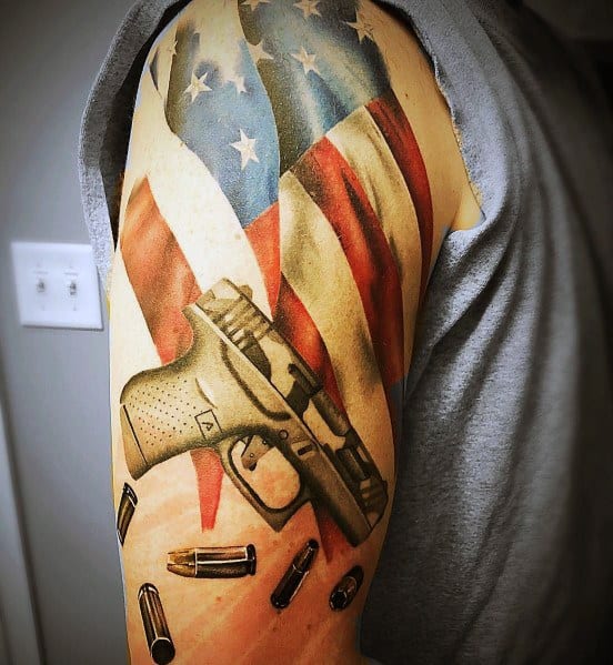 Color tattoo of a Glock pistol on the upper arm, featuring an American flag background and flying bullets, symbolizing patriotism and firearm culture.