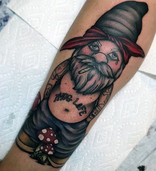 Tattoo of a gangster-style gnome with a black hat and red bandana, showing a "THUG LIFE" chest tattoo, gold tooth, and teardrop tattoo.