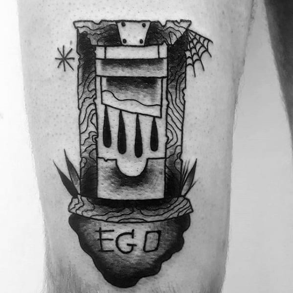 Black and grey guillotine tattoo with dripping blood, cobweb and star details, and the word “EGO” engraved on a stone base.