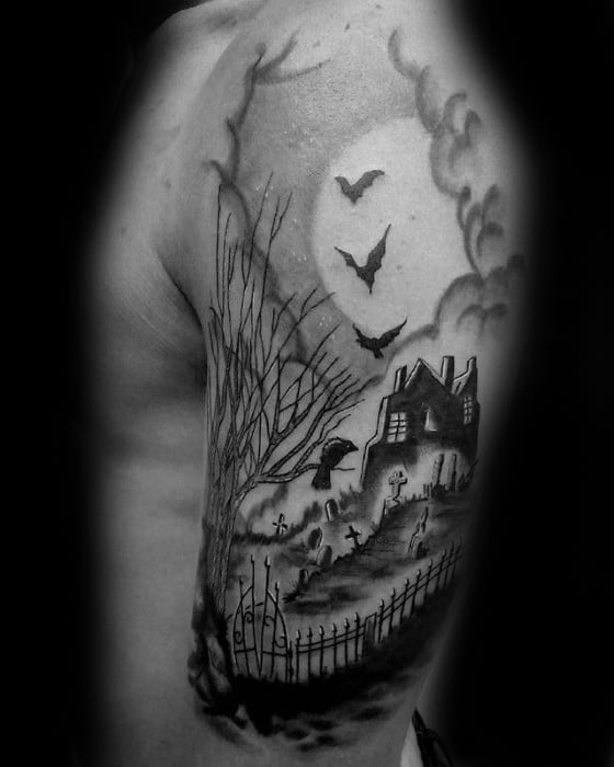 Upper arm tattoo of a haunted house and graveyard with flying bats, bare trees, wrought-iron gate, and a glowing full moon.