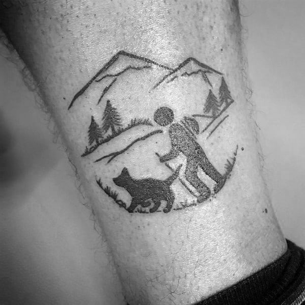 A black and gray minimalist tattoo of a hiker with a backpack and a dog on a trail, surrounded by mountains and trees, symbolizing adventure.