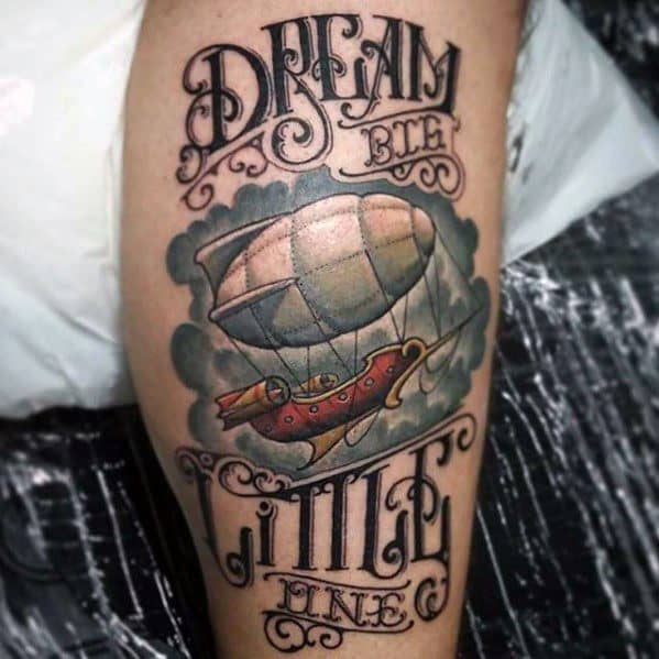 Colorful leg tattoo featuring a vintage airship with clouds, surrounded by ornate lettering that reads “Dream Big Little One.”