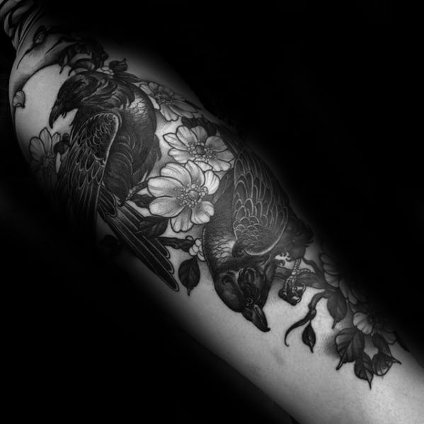 Black and grey tattoo of Huginn and Muninn surrounded by blooming flowers, inked on the forearm with fine detail and shading.