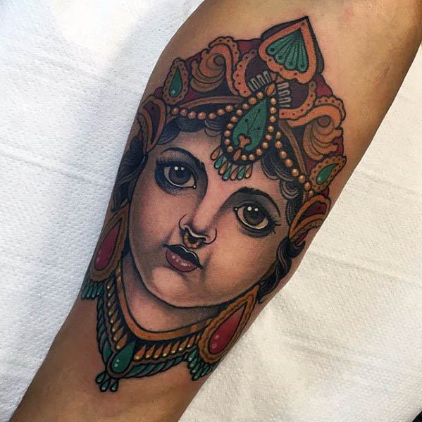 Colorful Krishna tattoo of a young Krishna's face adorned with traditional Indian jewelry, a crown, and expressive, wide eyes.