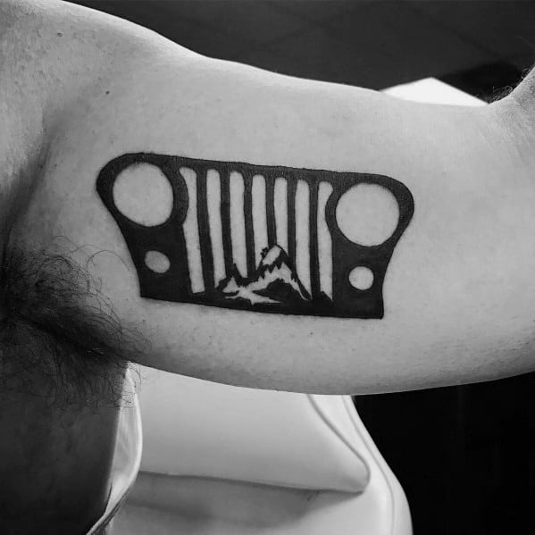 Minimalist black ink tattoo of a Jeep grille with a mountain silhouette inside, placed on the inner bicep.